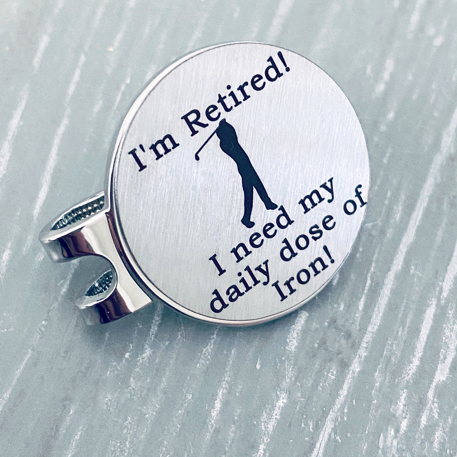 1 inch brushed silver stainless steel golf ball marker on magnetic hat clip. Marker is engraved with the image of a golfer swinging a club and the phrase "I'm retired. I need my daily dose of Iron!"