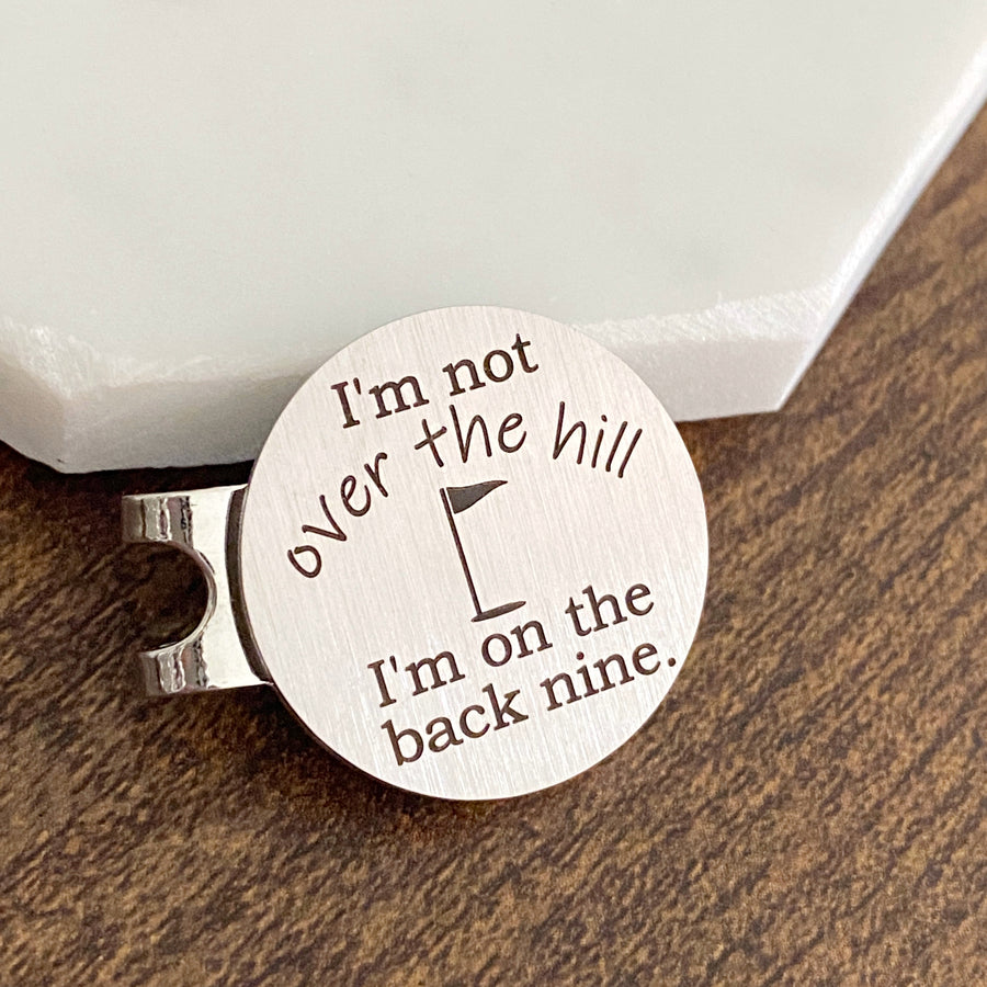 1 inch brush metal magnetic golf ball marker and hat clip engraved with "I'm not over the hill. I'm on the back nine." with the image of a golf flag pole