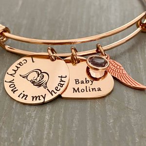 rose gold bangle charm bracelet with a 3/4" round engraved disc with the image of a baby sleeping under angel wings and the verbiage "I carry you in my heart" next to it is a 3/4" heart engraved with the name "Baby Molina" and. a diamond april birthstone. Last is an angel wing charm.