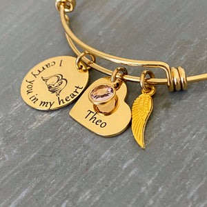 yellow gold bangle charm bracelet with a 3/4" round engraved disc with the image of a baby sleeping under angel wings and the verbiage "I carry you in my heart" next to it is a 3/4" heart engraved with the name "Theor" and a purple June birthstone. Last is an angel wing charm.