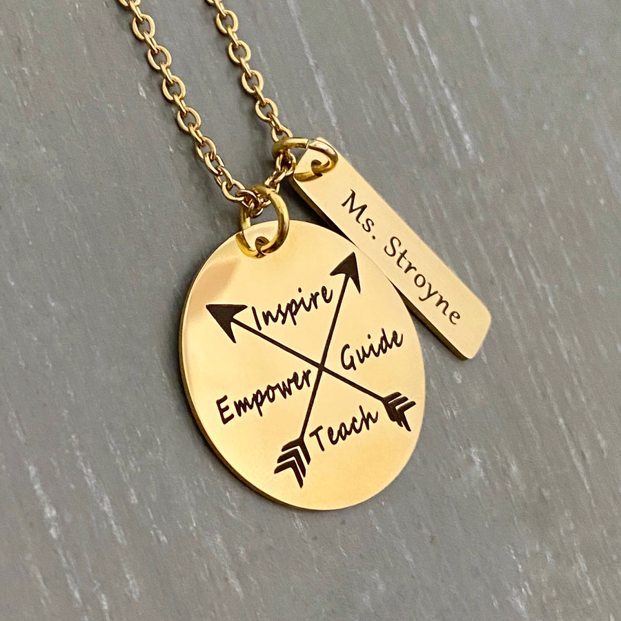 A 1 inch plated yellow gold disc engraved with a cross arrow design and the words Inspire, Empower, Guide, and Teach. Next to the disc is a 1.2" rectangle with the teachers name "Ms. Stroyne". Charms are attached to a yellow gold cable chain