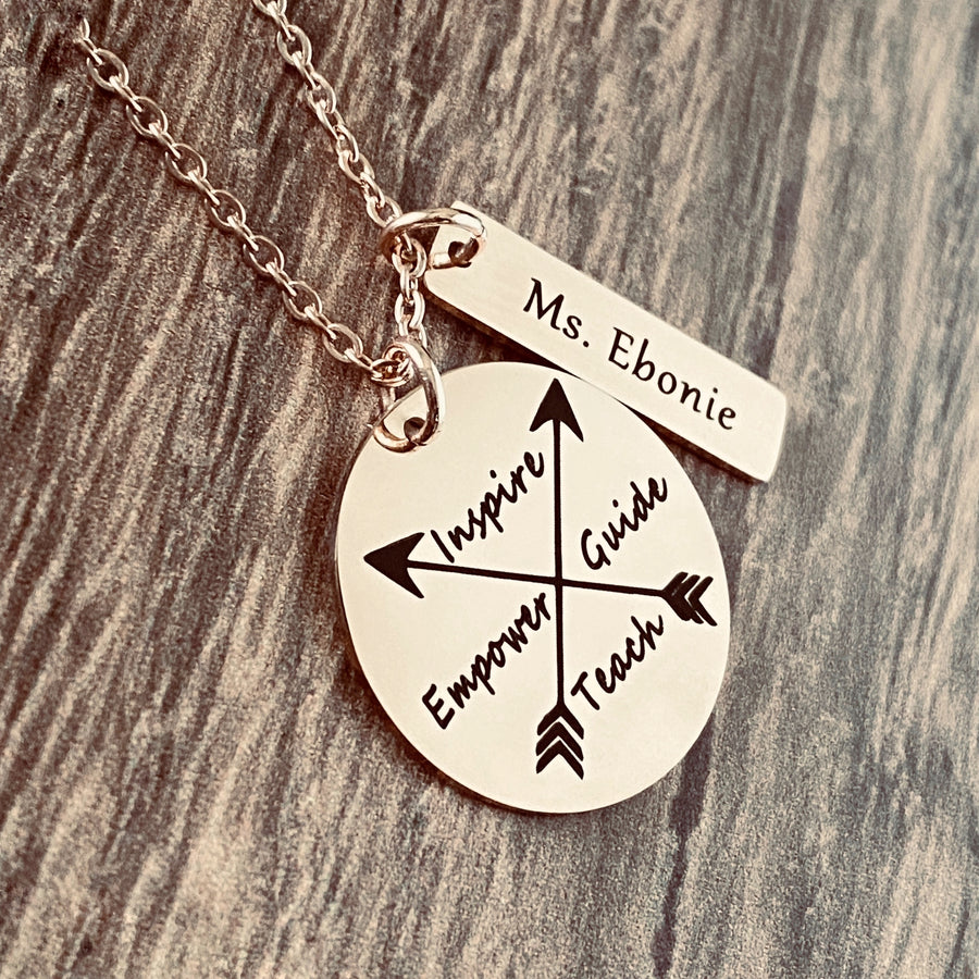 A 1 inch plated rose gold disc engraved with a cross arrow design and the words Inspire, Empower, Guide, and Teach. Next to the disc is a 1.2" rectangle with the teachers name "Mrs. Barrio". Charms are attached to a rose gold cable chain