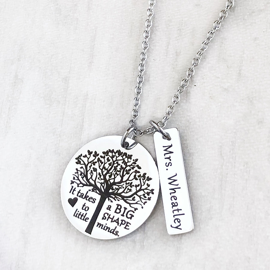 A 1-inch round stainless steel silver disc engraved with a tree of life symbol and the verbiage "it takes a BIG heart to shape little minds." Next to the disc is attached a 1-inch rectangle tag engraved with the name "Mrs. Wheatly". Both Charms are attached to a silver cable chain