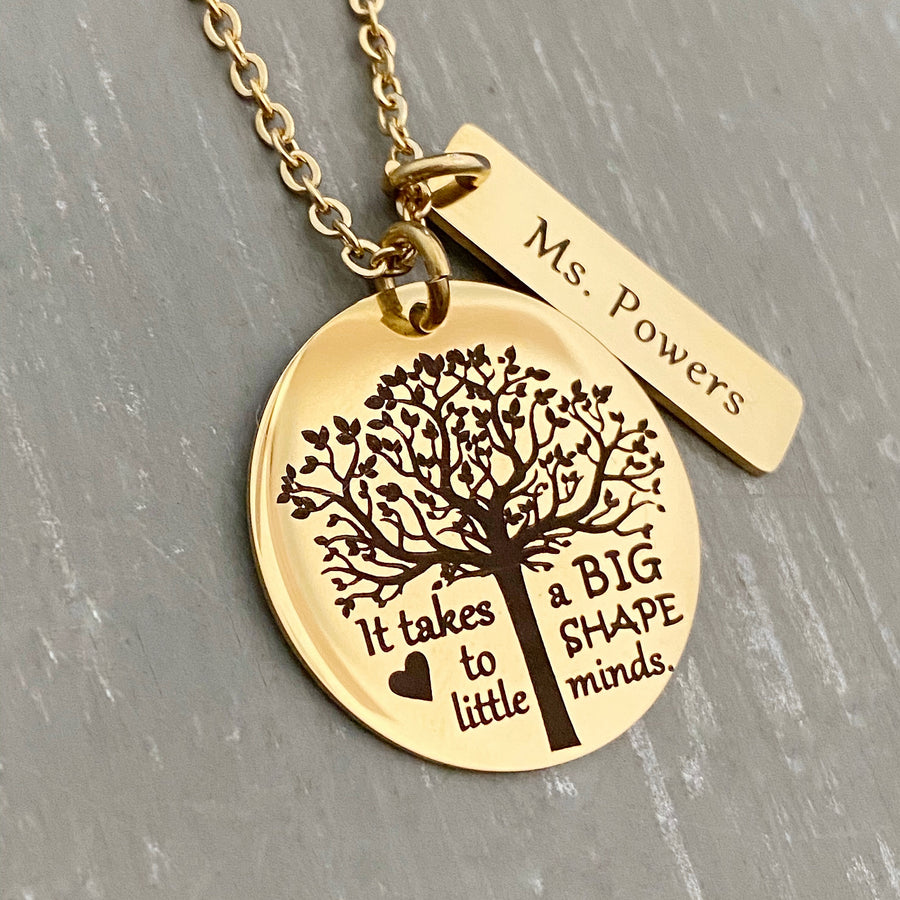 A 1-inch round stainless steel plated yellow gold disc engraved with a tree of life symbol and the verbiage "it takes a BIG heart to shape little minds." Next to the disc is attached a 1-inch rectangle tag engraved with the name "Ms Powers". Both Charms are attached to a yellow gold cable chain