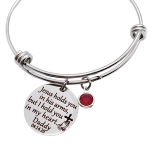 Silver bangle charm bracelet with 7/8" engraved disc with the verbiage "jesus holds you in his arms, but i hold you in my heart." with the name "mom" and date "7.30.12" as well as the image of jesus playing hands holding a cross. Next to the disc is a red january crystal stone