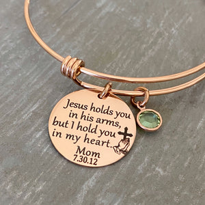 Rose gold bangle charm bracelet with 7/8" engraved disc with the verbiage "jesus holds you in his arms, but i hold you in my heart." with the name "mom" and date "7.30.12" as well as the image of jesus playing hands holding a cross. Next to the disc is a august crystal stone