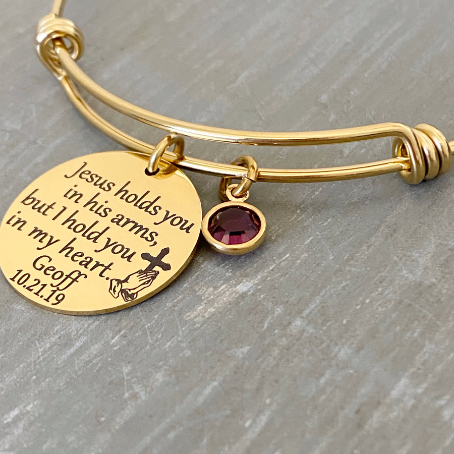 yellow gold bangle charm bracelet with 7/8" engraved disc with the verbiage "jesus holds you in his arms, but i hold you in my heart." with the name "mom" and date "7.30.12" as well as the image of jesus playing hands holding a cross. Next to the disc is a purple february stone