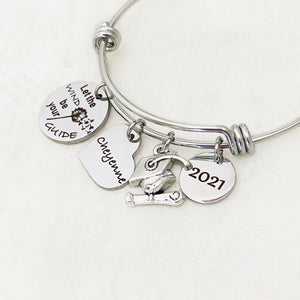 silver stainless steel bangle charm bracelet with a 3/4 inch charm engraved with a dandelion image and "let the wind be your guide", a 3/4 inch personalized heart name charm, graduation hat charm, and a .5 inch "2021" year charm.