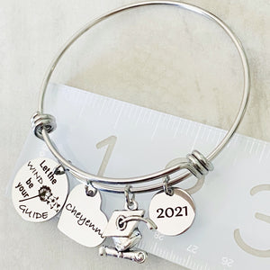 silver stainless steel bangle charm bracelet with a 3/4 inch charm engraved with a dandelion image and "let the wind be your guide", a 3/4 inch personalized heart name charm, graduation hat charm, and a .5 inch "2021" year charm. shown on a ruler