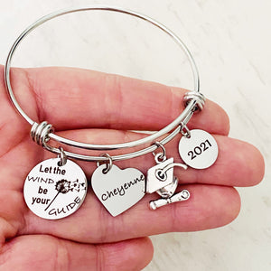 silver stainless steel bangle charm bracelet with a 3/4 inch charm engraved with a dandelion image and "let the wind be your guide", a 3/4 inch personalized heart name charm, graduation hat charm, and a .5 inch "2021" year charm. shown on a hand. shown in palm
