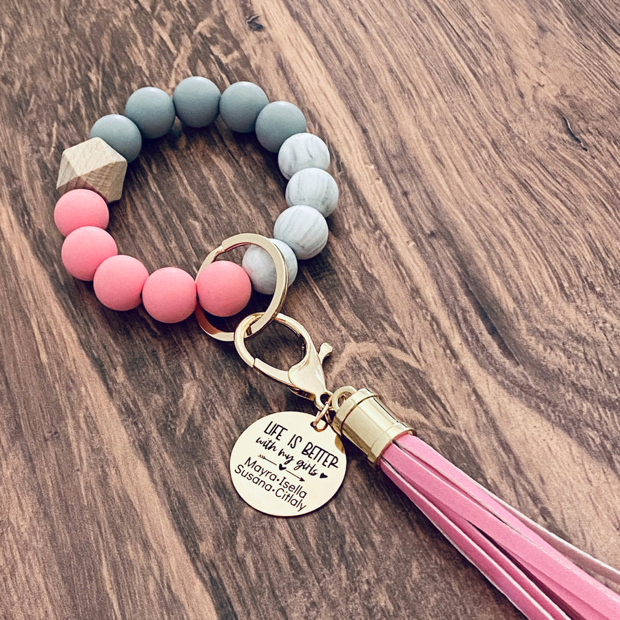 Pink, white, grey silicone beaded bracelet with leather lobster hook keychain tassel. Engraved round charm tag with "Life is better with my girls" a heart arrow design, and personalized names "Mayra, Isella, Susana, Citaly""