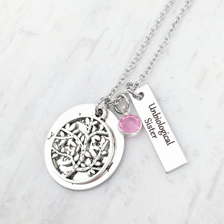 Round pendant necklace engraved with the phrase "like roots to a tree you are connected to my heart". a tree of life charm lays on top of the engraved round pendant. Next to the pendant is a rectangle charm engraved with "unbiological sisters". attached to the main pendant is a october birthstone. all charms are attached to a stainless steel silver cable chain.