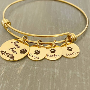 Yellow Gold Bangle adjustable charm bracelet with a 3/4" engraved disc with a bear paw print, mountain range scene and verbiage "Mama Bear" along with three 1/2" name tags engraved with a cub paw print and the names Gavyn, Harlyn, Scotlyn