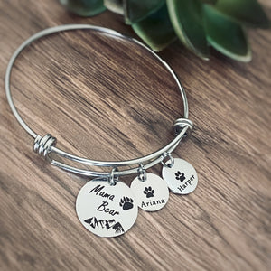 Silver Bangle adjustable charm bracelet with a 3/4" engraved disc with a bear paw print, mountain range scene and verbiage "Mama Bear" along with three 1/2" name tags engraved with a cub paw print and the names Ariana and Harper
