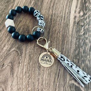 Cow print and black round silicone beaded wristlet with large lobster hook leather tassel that matches. A rose gold charm attached engraved with "mama mode. All Day Every Day" with boho rainbow image 