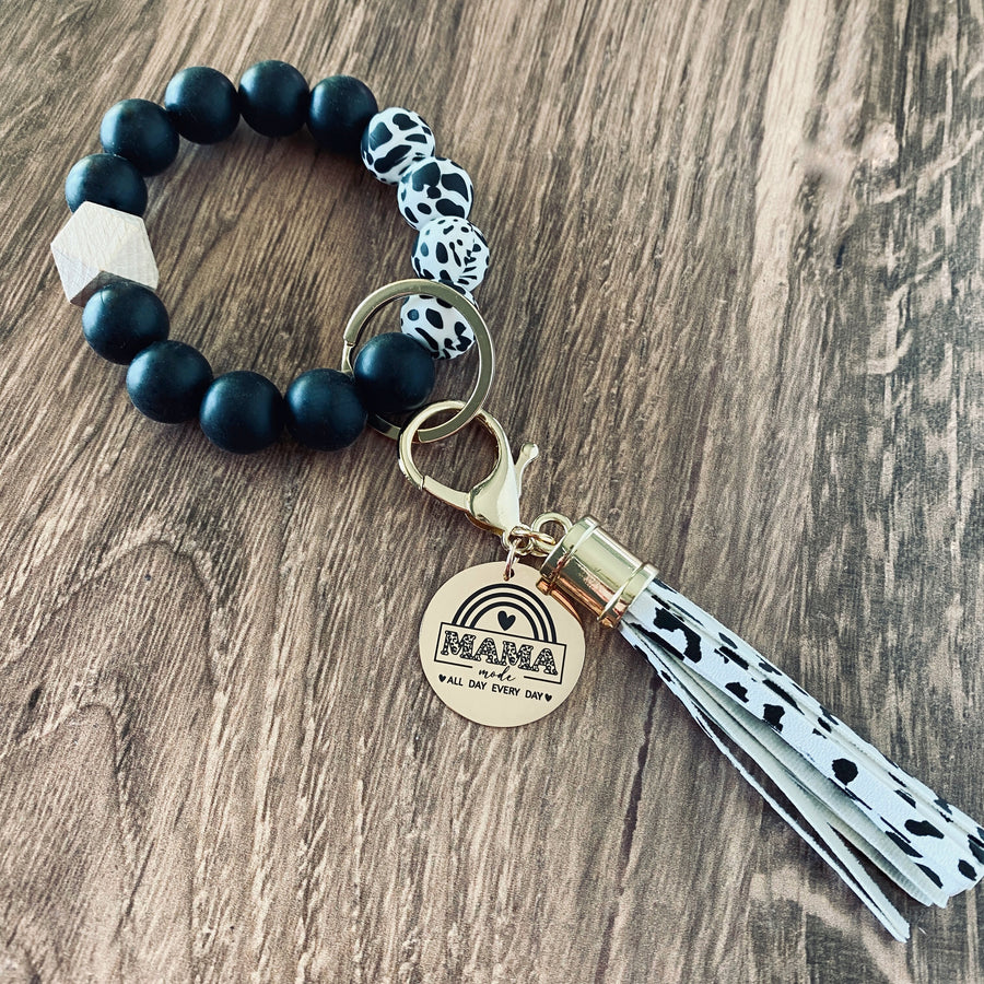 Cow print and black round silicone beaded wristlet with large lobster hook leather tassel that matches. A rose gold charm attached engraved with "mama mode. All Day Every Day" with boho rainbow image 