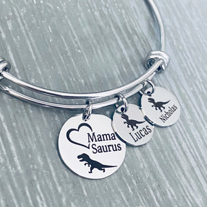silver stainless steel triple loop bangle bracelet. first round charm measures .75 inches and is engraved with a trex dinosaur image and on open heart frame with "Mama Suarus". Next charm measures 1/2 inch and is engraved with a baby trex and the name lucas. next charm is engraved with a baby trex and the name nicholas