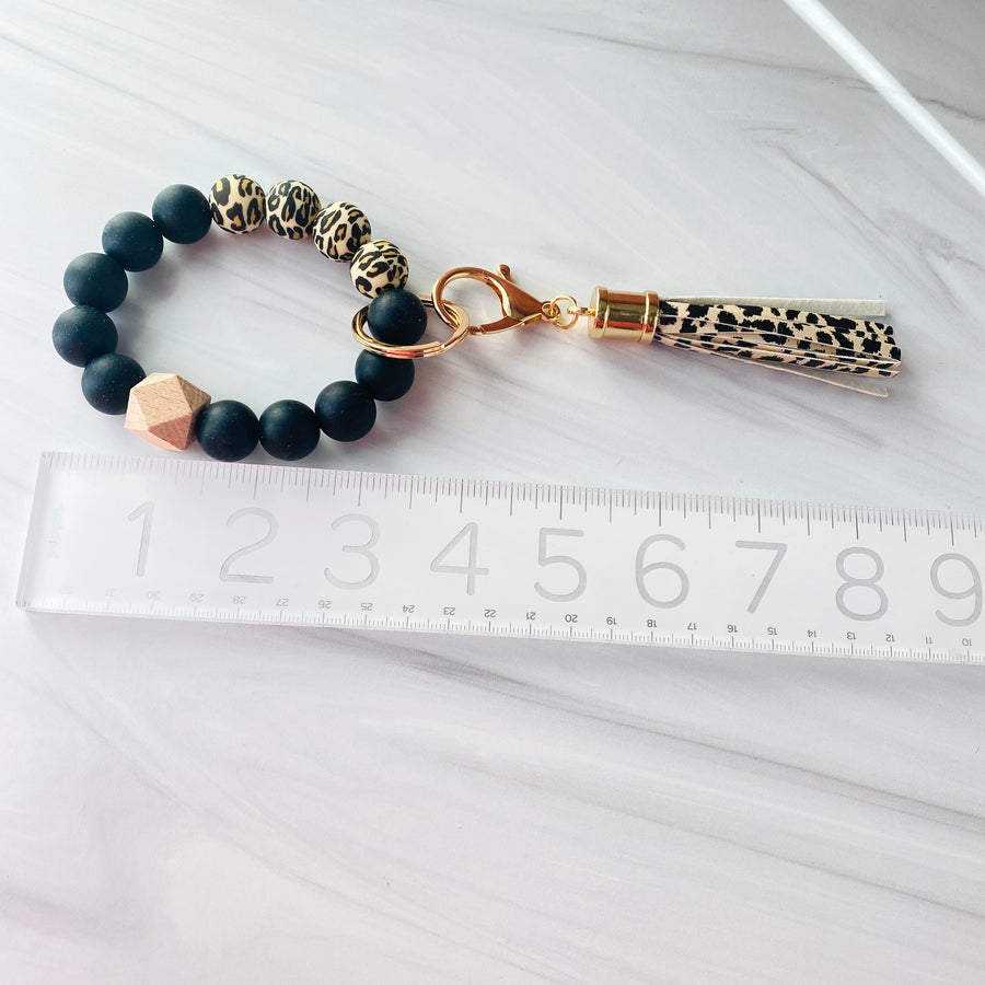 Cheetah print and black silicone beaded wristlet bracelet with one wood bead and a cheetah tassel hooked to the bracelet against a ruler to show 9" long including the tassel dangling.