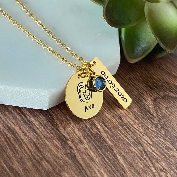stainless steel yellow gold round pendant engraved with an image of a mother holding a baby and the name Ava. Next to it is a 1 inch rectangle with the birthdate 09.09.2020 and a blue september birthstone. the charms are attached to a cable chain