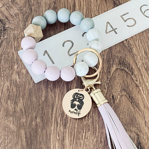 purple, white, grey round silicone beaded wristlet with matching leather tassel keychain. attached is a messy bun mom image and the inscription "#momlife". bracelet on ruler to show measurements