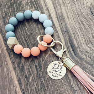 coral, white, grey silicone beaded bracelet with leather lobster hook keychain tassel. Engraved round charm tag with "Mom of Girls" a heart arrow design, and personalized names "Maryn, Peyton, Baylea"