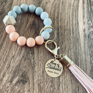 pink, white, and grey round silicone beaded wristlet with large lobster hook leather tassel that matches. A rose gold charm attached engraved with "mom of sassholes" and personalized with the names "Harper and Henry"