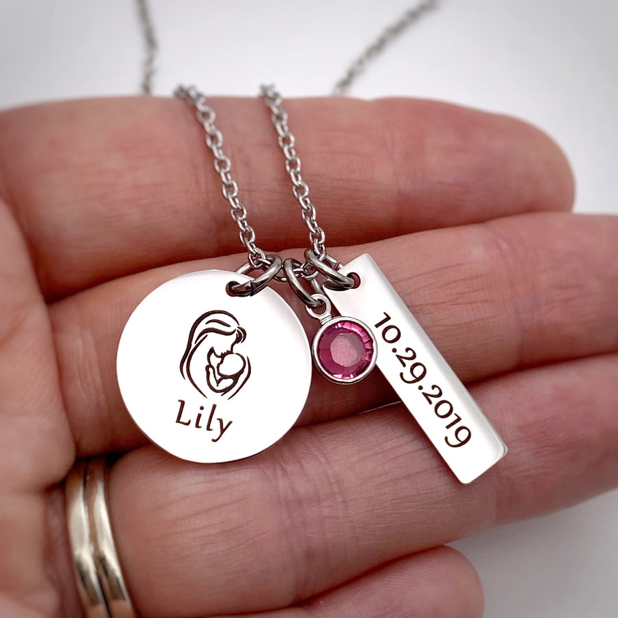 stainless steel silver round pendant engraved with an image of a mother holding a baby and the name Lily. Next to it is a 1 inch rectangle with the birthdate 10.29.2019 and a pink october birthstone. the charms are attached to a cable chain on a womens hand