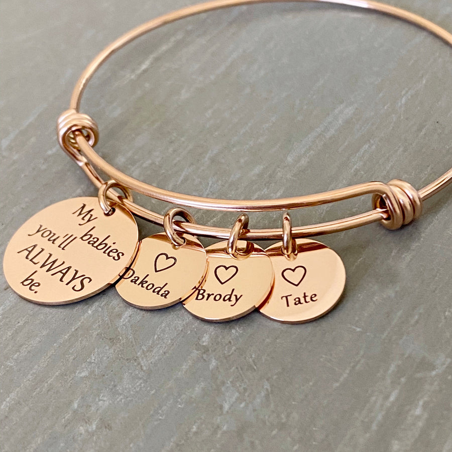 Rose gold bangle charm bracelet with a 3/4" disc engraved with "my babies you'll always be." and a 1/2" engraved disc with an open heart and the name Dakoda, a second charm disc with the name Brody, and a third name disc with the name tate.