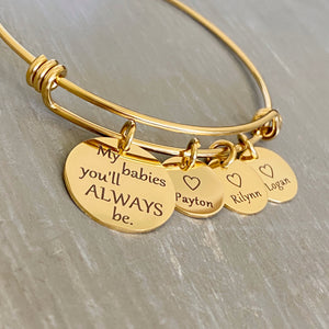 Yellow gold bangle charm bracelet with a 3/4" disc engraved with "my babies you'll always be." and a 1/2" engraved disc with an open heart and the name Payton, a second charm disc with the name Rilynn, and a third name disc with the name Logan.