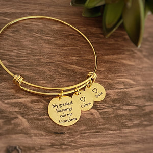 stainless steel yellow gold bangle charm bracelet. first charm is engraved with the saying "my greatest blessings call me Grandma" next is a small 1/2" charm tag engraved with a heart and the name Cohen. Next is a 1/2" charm engraved with a heart and the names duke