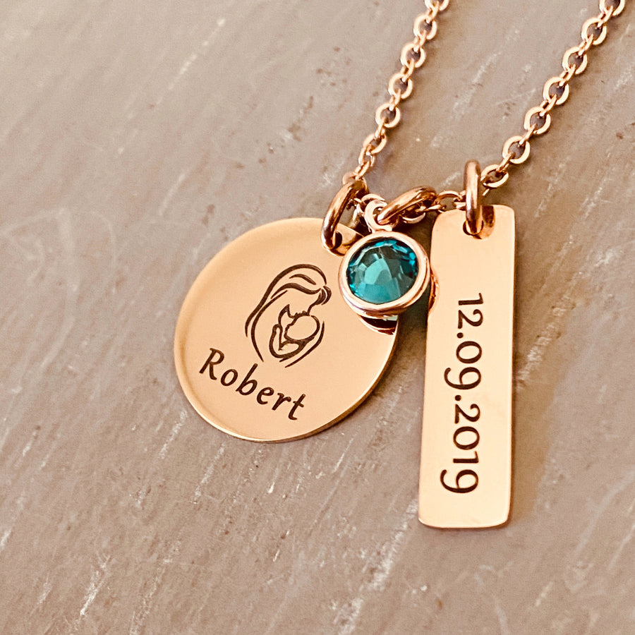 stainless steel plated rose gold round pendant engraved with an image of a mother holding a baby and the name Robert. Next to it is a 1 inch rectangle with the birthdate 12.09.2019 and a blue december birthstone. the charms are attached to a cable chain