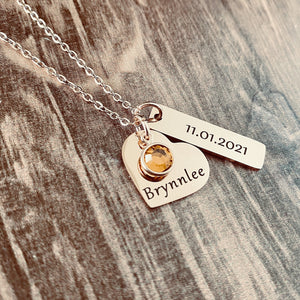stainless steel rose gold necklace with a 3/4 inch engraved heart with the name Brynnlee and a november orange birthstone. Next to the heart hangs a 1" rectangle charm with the date of birth 11.01.21