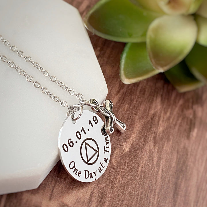 Round silver 7/8" pendant charm necklace engraved with "One Day at a Time", the AA sobriety symbol and the recovery date of 06.01.19. Next to the round charm is a silver religious cross charm. Both pieces are attached to a stainless steel cable chain 