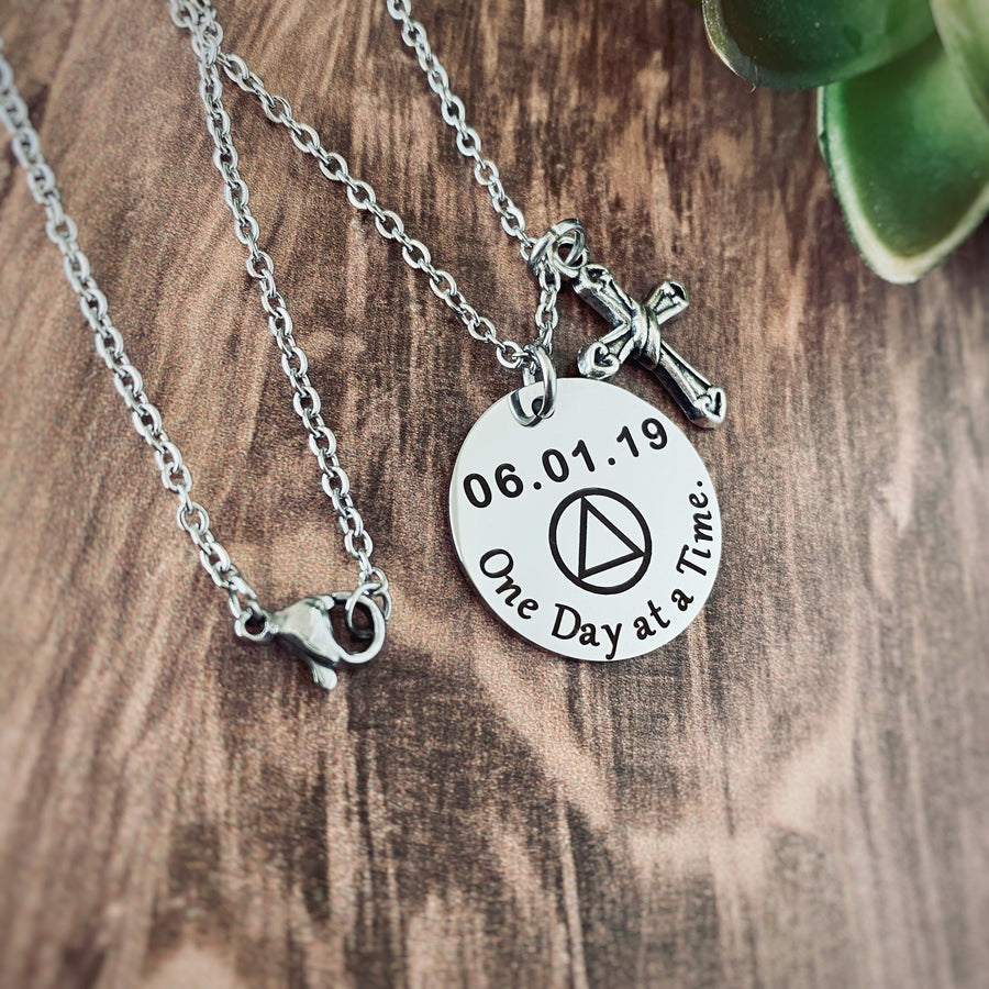 Round silver 7/8" pendant charm necklace engraved with "One Day at a Time", the AA sobriety symbol and the recovery date of 06.01.19. Next to the round charm is a silver religious cross charm. Both pieces are attached to a stainless steel cable chain with lobster clasp