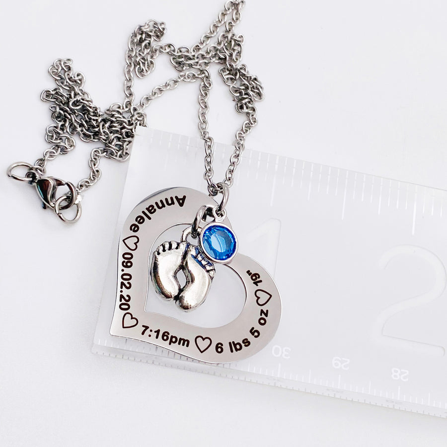 Silver Stainless Steel Open heart pendant attached to a silver cable chain. Baby feet charm and september birthstone attached. Black engraving around the heart charm with the name annalee, birthdate 09.20.20, time of birth 7:16pm, weight 6 lbs 5 oz, and length of 19" on ruler showing 1.5" wide
