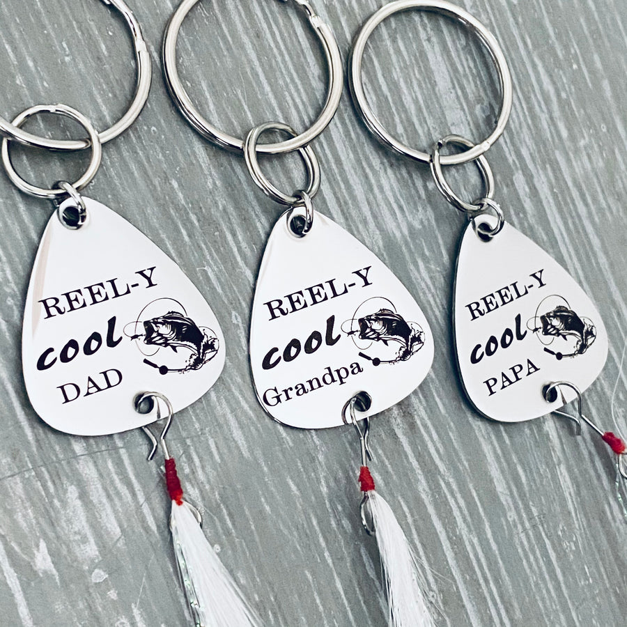 Silver stainless steel fishing lure keychain showing 3 options, REEL-Y cool Dad, Grandpa, or Papa. Each keychain will also be engraved with a bass fish with fishing line. Attached on the bottom of the lure is a fishing feather. The top is attached to a triple loop keyring