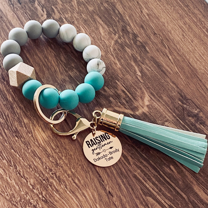 Turquoise round silicone beaded bracelet with rose gold charm tag engraved with "raising gentlemen" heart arrow, and boys names dakoda, brody, and tate