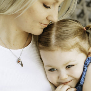 mother holding daughter while wearing heart charm name necklace with rectangle date charm tag