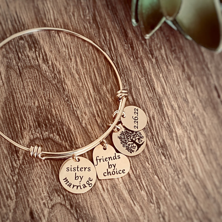 rose gold Sister in Law  charm bracelet with 4 charms. First charm is engraved with "sister by marriage". next is a tree of life symbol char. next is a heart charm engraved with "friends by choice" last charm is a round charm engraved with date 2.26.22