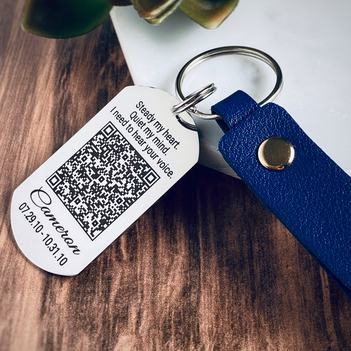 silver stainless steel dog tag keychain attached to a blue  engraved with the phrase Your voice is my favorite sound. Then a voicemail QR code. Underneath the qr code is the name Cameron, date of birth 5.31.62, and death date 4.08.19.