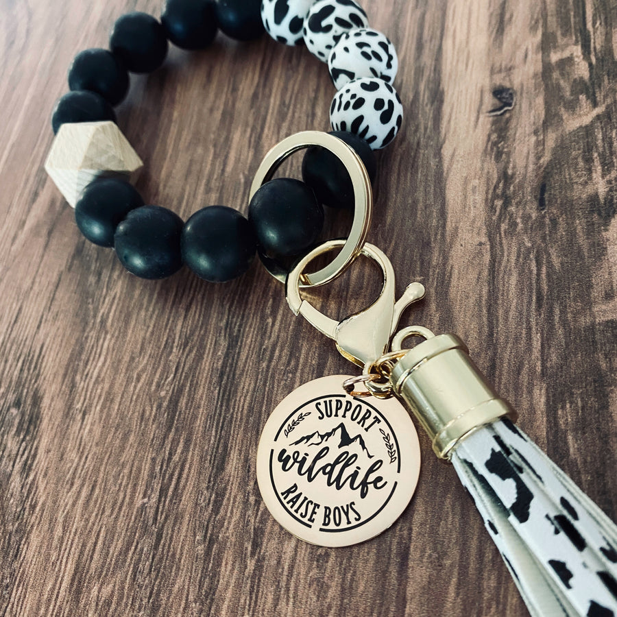 cow print wristlet with keychain leather tassel and charm tag engraved with "support wildlife raise boys"