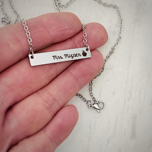 Silver stainless steel horizontal  bar necklace engraved with "mrs. Mizner" and an apple charm. attached to a stainless steel cable chain pictured on a hand