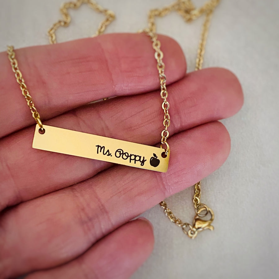 Yellow Gold stainless steel horizontal  bar necklace engraved with "Mrs. Poppy" and an apple charm. attached to a stainless steel cable chain on a hand