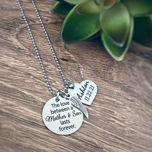 Silver 7/8" engraved round pendant memorial necklace with "The love between a mother & son lasts forever". Attached next is an angel wing charm. after that dangles a 3/4" heart charm engraved with the name Ashton and the death date 11.22.21 All grief charms are connected to a stainless steel cable chain with lobster clasp. 