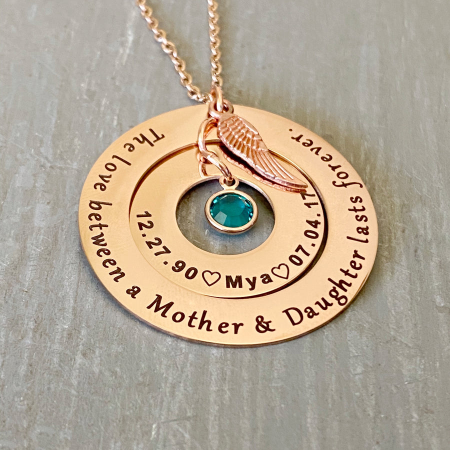 Rose Gold Round 2 disc necklace. outside washer is engraved with "the love between a mother and daughter lasts forever." The inner washer is engraved with the birthdate 12.27.90 a heart image the name Mya, heart image, and death date 7..04.17. May birthstone dangles inside the washer and angel wing charm on top. All charms and pendants hang from a stainless steel cable chain