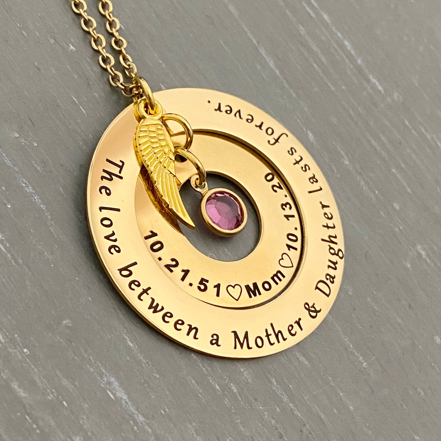 Yellow Gold Round 2 disc necklace. outside washer is engraved with "the love between a mother and daughter lasts forever." The inner washer is engraved with the birthdate 10.21.51 a heart image the name Mom, heart image, and death date 10.13.20. October pink birthstone dangles inside the washer and angel wing charm on top. All charms and pendants hang from a stainless steel cable chain