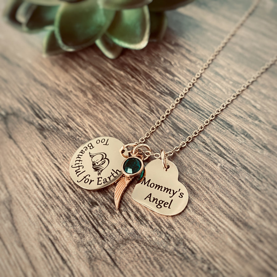 rose gold round disc engraved with too beautiful for earth and an image of a baby with angel wings, an angel wing, a heart engraved with the name nora and a green may stone. Pendant charms are attached to a rose gold cable chain.