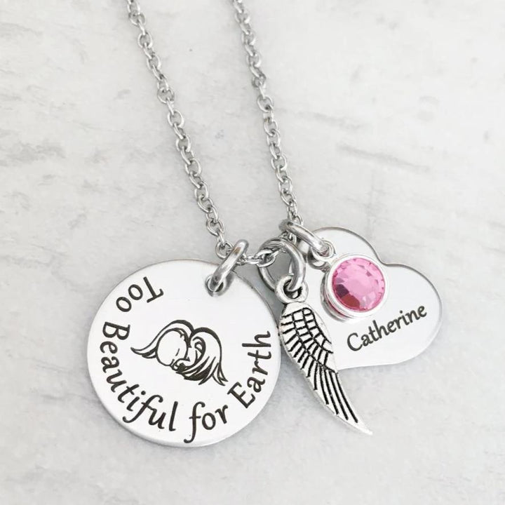 silver round disc engraved with too beautiful for earth and an image of a baby with angel wings, an angel wing, a heart engraved with the name Catherine and a pink October stone. Pendant charms are attached to a silver cable chain.