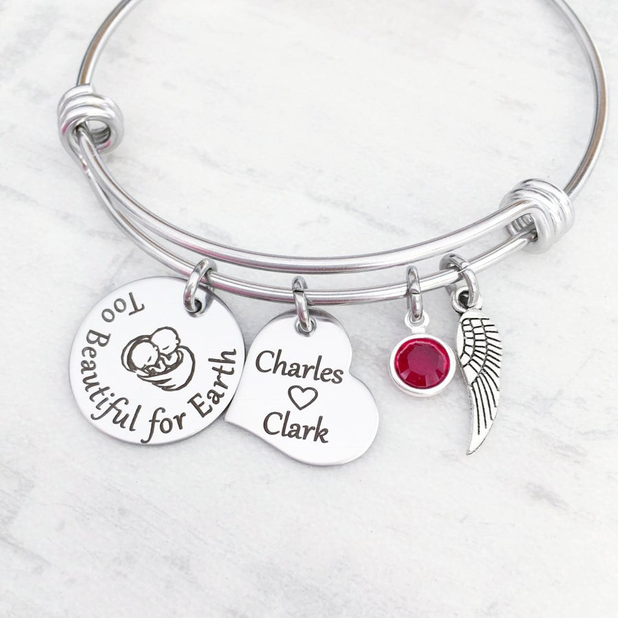 Silver stainless steel trip loop expandable bangle charm bracelet. first charm is engraved with twin babies under angel wings along with the saying "Too beautiful for earth". next is a heart charm engraved with the names charles and clark. next is the twins birthstones and an angel wing charm.