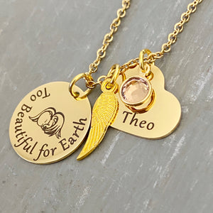 Yellow Gold round disc engraved with too beautiful for earth and an image of a baby with angel wings, an angel wing, a heart engraved with the name Theo and a clear april stone. Pendant charms are attached to a yellow gold cable chain.